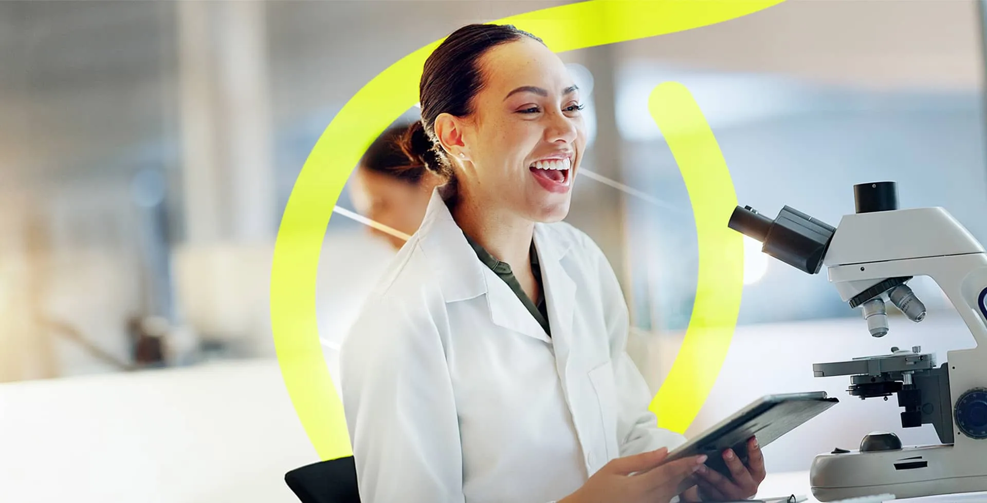 A smiling woman in a lab coat laughing next to a microscope in a bright laboratory setting.