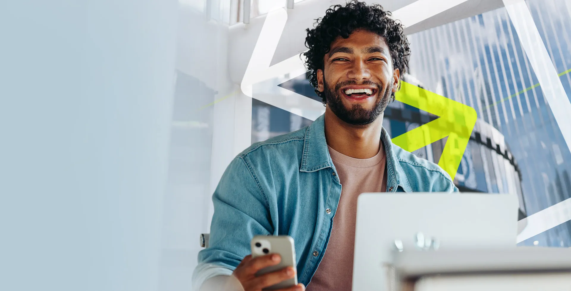 man sat in an office with a laptop in front of him and a phone in his hand as he smiles at the camera, happy exploring Marketing, Sales and Comms roles on TalentNext.