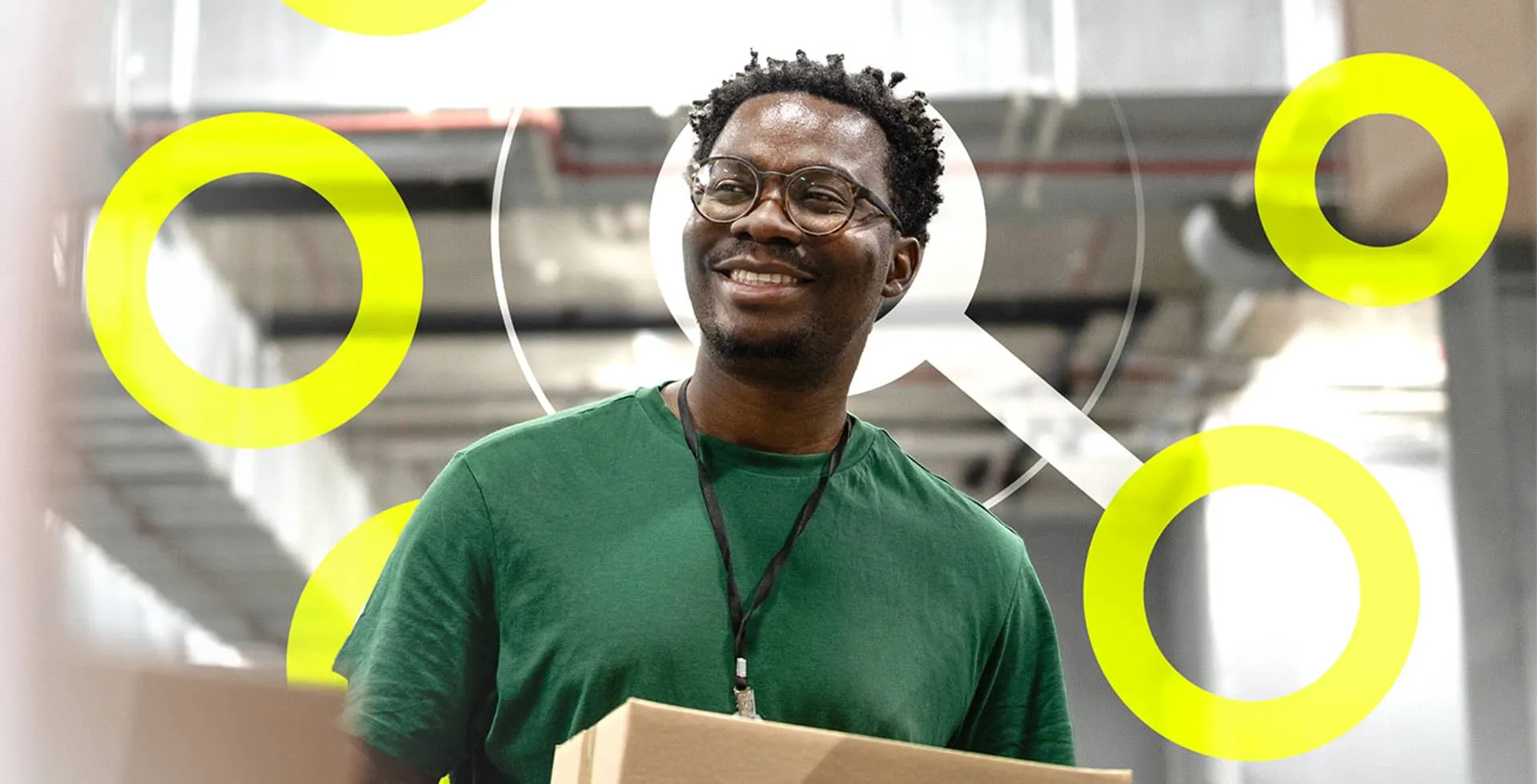 A smiling man in a green t-shirt and glasses holding a parcel in a warehouse setting.