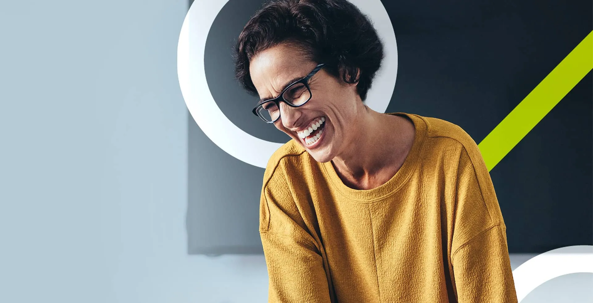 woman wearing glasses sat in an office with a dark screen in the background, laughing as she looks down to her left, excited about finding an opportunity in Finance on TalentNext.