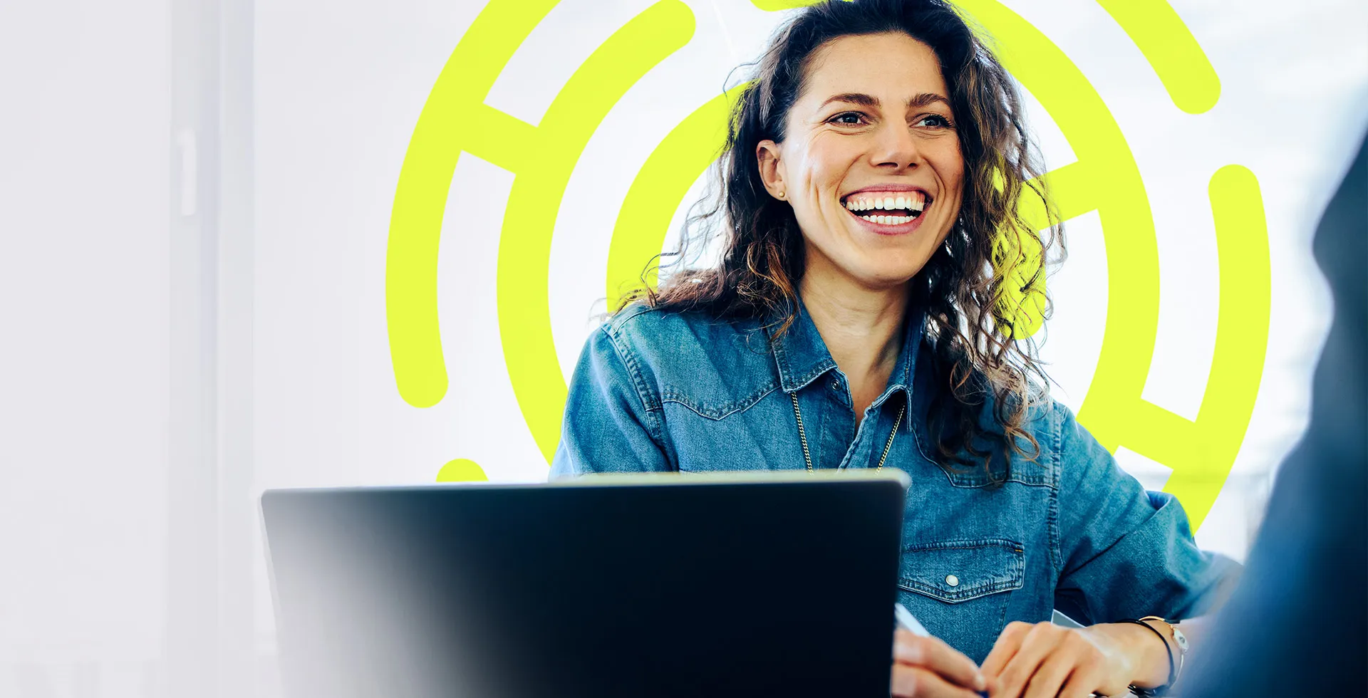 woman with long wavy hair sat in front of a laptop as she smiles and interacts with a colleague, happy in the insurance role she found on TalentNext.